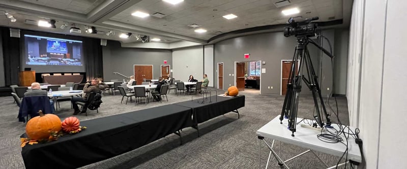 Large room with venue space for people at tables, cameras on a stand, a stage, and a projected screen